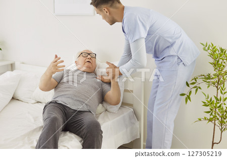 Patient young caregiver assisting indignant frustrated elderly man who refusing to settle into bed. Patient young caregiver assisting indignant frustrated elderly man who refusing to settle into bed. 127305219
