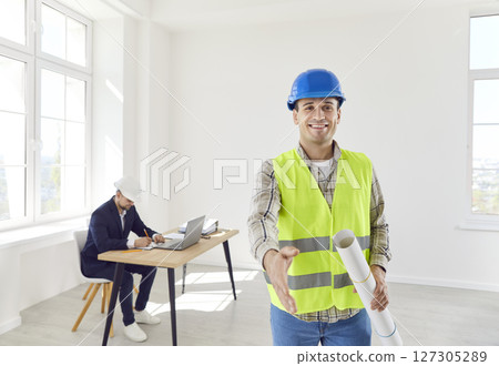 Foreman shaking hand to camera making deal holding blueprint with architect working in background. 127305289
