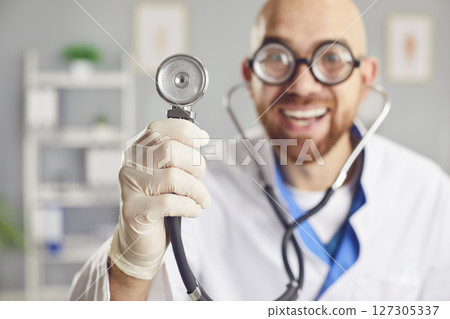 Portrait of a young funny smiling doctor in medical uniform standing with stethoscope in clinic. 127305337