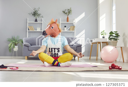 Funny man in sportswear and horse head mask doing yoga exercise sitting on the floor in lotus pose. 127305371