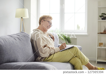 Mature woman sitting on sofa in living room at home and writing notes in her notepad. 127305394