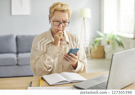 Pensive woman wearing glasses sitting at desk with laptop and phone, thinking about task or message. Pensive woman wearing glasses sitting at desk with laptop and phone, thinking about task or message. 127305399