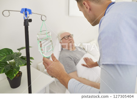 Caring young male nurse checking on elderly patient during IV infusion, providing support in clinic. Caring young male nurse checking on elderly patient during IV infusion, providing support in clinic. 127305404