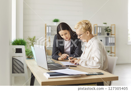 Two focused female colleagues sitting in modern office, reviewing data on sheet, working as team. Two focused female colleagues sitting in modern office, reviewing data on sheet, working as team. 127305405