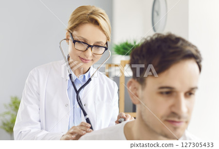 Female doctor carefully examines and listens to lungs of male patient with stethoscope. 127305433
