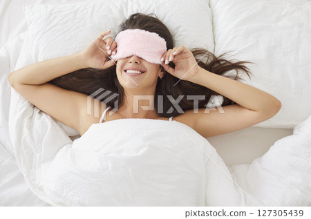 Top view portrait of a happy young woman wearing sleep face mask lying on bed in the morning Top view portrait of a happy young woman wearing sleep face mask lying on bed in the morning 127305439