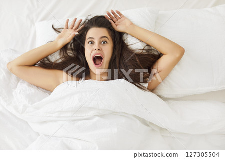 Frightened young woman lying in bed, shouting in panic, realizing she overslept and late for work. 127305504