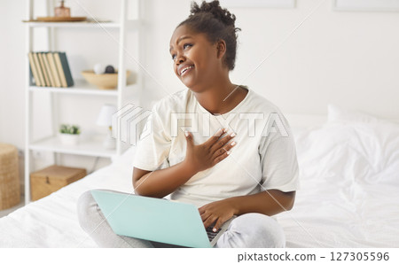 Young African American woman sitting on bed with laptop, smiling skeptically at something amusing. 127305596