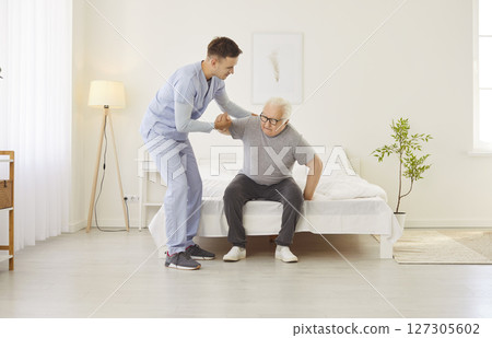 Portrait of a young friendly male nurse helping a senior patient to get off the bed at home. 127305602