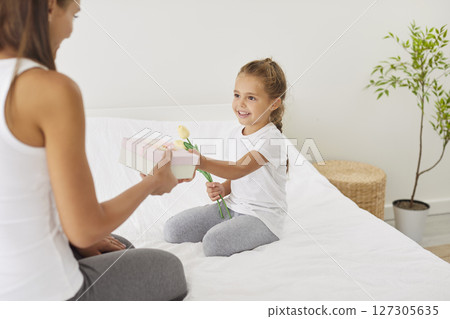 Smiling girl sitting with mother on bed, giving her gift box and flowers for Mother Day or birthday. 127305635
