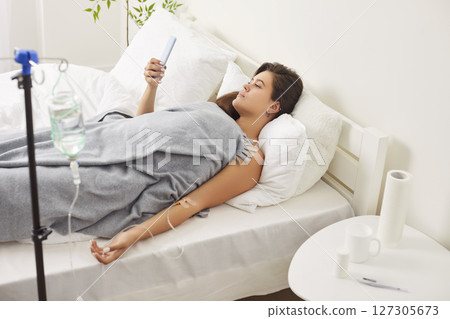 Sick young woman lying on bed in bedroom at home receiving IV drip infusion using mobile phone. Sick young woman lying on bed in bedroom at home receiving IV drip infusion using mobile phone. 127305673