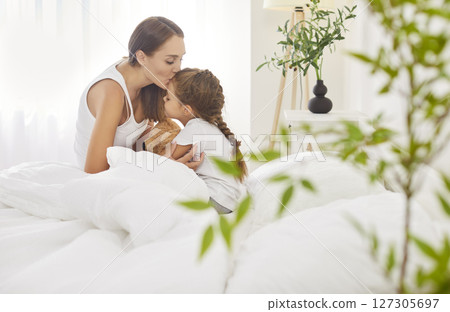 Loving mother embracing her daughter sitting in bed, kissing her on forehead with love and care. 127305697