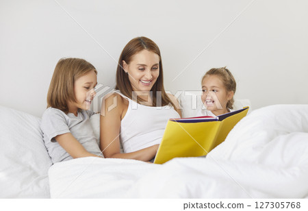 Young happy mother reading a book to her children sitting on bed in bedroom before going to sleep 127305768