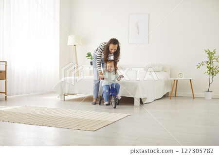 Young mother teaching happy cute child girl to ride bike in living room at home. 127305782