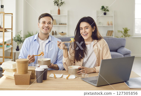 Happy couple sitting at table with laptop and eating fresh fast food. Delivery service concept. 127305856