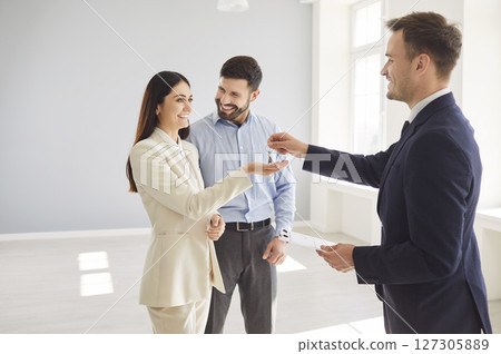 Real estate agent handing property keys to happy smiling young family couple in bright empty room. Real estate agent handing property keys to happy smiling young family couple in bright empty room. 127305889