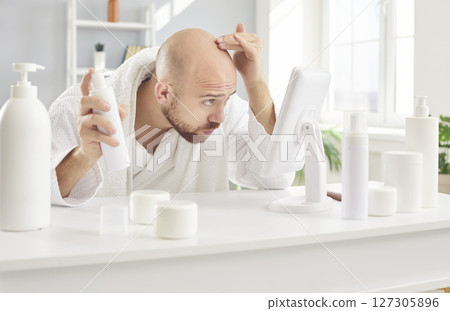 Balding young man in bathrobe looking in mirror and applying hair growth lotion on his scalp Balding young man in bathrobe looking in mirror and applying hair growth lotion on his scalp 127305896