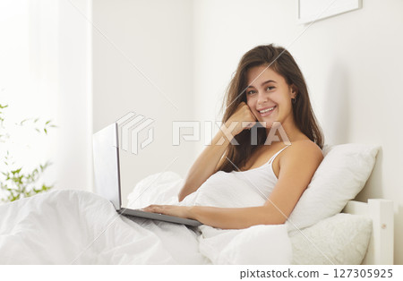 Young woman sitting on bed in bedroom in morning with laptop looking at camera and smiling 127305925