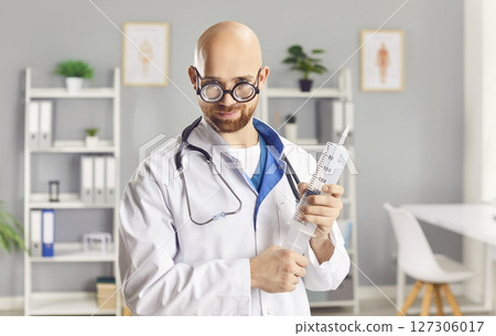 Funny young man doctor holding big syringe standing in medical clinic. 127306017