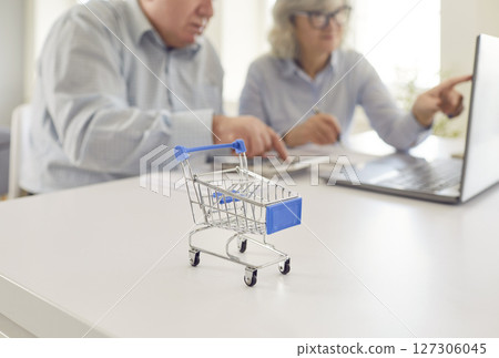 Senior couple calculating finances or planning purchases with a shopping cart in the foreground. 127306045