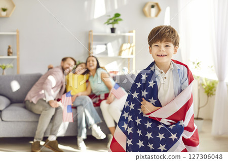 Child boy wrapped in us flag looking at camera with his happy american family in background. Child boy wrapped in us flag looking at camera with his happy american family in background. 127306048