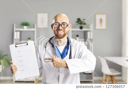 Funny man doctor pointing to report file with appointment and looking at camera in medical clinic Funny man doctor pointing to report file with appointment and looking at camera in medical clinic 127306057