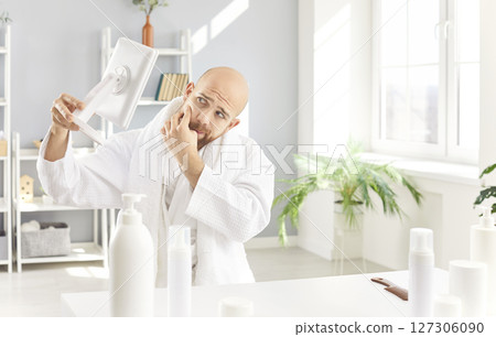 Funny man relaxing in white bathrobe with bald head looking in mirror doing spa pedicure at home. Funny man relaxing in white bathrobe with bald head looking in mirror doing spa pedicure at home. 127306090