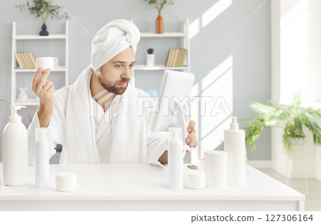 Funny man in white bathrobe and towel sitting at table with cosmetic bottles having beauty day 127306164