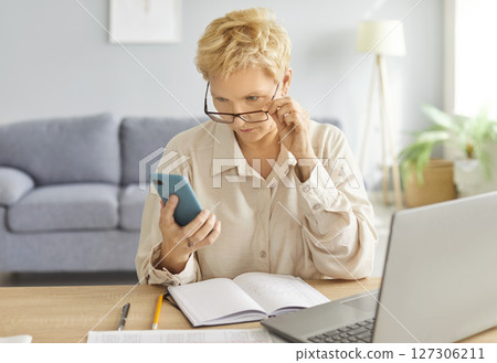 Blonde woman sitting at desk in living room focused on smartphone, reading important work message. Blonde woman sitting at desk in living room focused on smartphone, reading important work message. 127306211