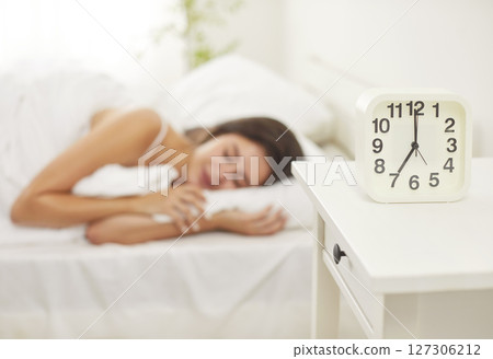 Young peaceful woman sleeping on bed under white blanket at home with alarm clock on table. Young peaceful woman sleeping on bed under white blanket at home with alarm clock on table. 127306212