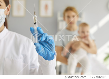 Closeup nurse holding syringe with vaccine, with mother and baby in blurred background 127306408