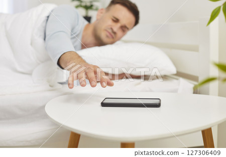 Young sleepy man waking up in the morning and stretching to mobile phone lying in bed at home. Young sleepy man waking up in the morning and stretching to mobile phone lying in bed at home. 127306409