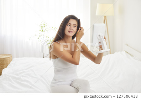 Young woman sitting on bed with mirror, brushing her hair with wooden comb during morning routine. 127306418