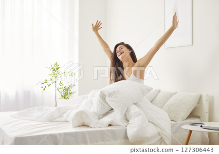 Happy woman sitting in bed under blanket, stretching after waking up, enjoying start of new day. 127306443
