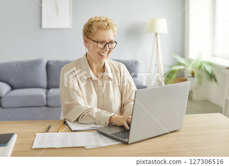 Happy smiling mature woman sitting on workplace at the desk working on laptop at home. 127306516