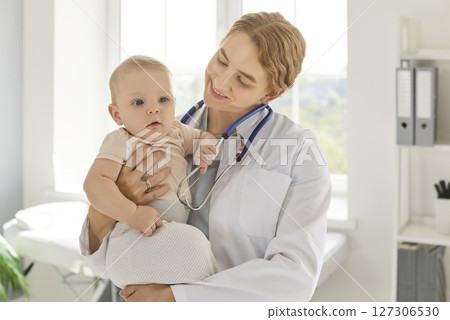 Caring smiling female pediatrician holding newborn toddler baby during health checkup in clinic. 127306530