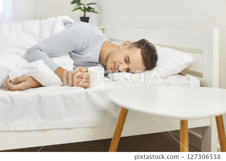 Young man sleeping well in bed on soft white pillow holding mug, resting, good night sleep Young man sleeping well in bed on soft white pillow holding mug, resting, good night sleep 127306538