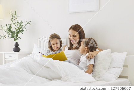Mother lies in bed with her two children in morning and reads book looking at illustrations. 127306607