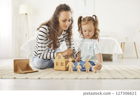 Little girl playing with wooden house and toy figures on carpet with mother in sunlit bedroom. Little girl playing with wooden house and toy figures on carpet with mother in sunlit bedroom. 127306621