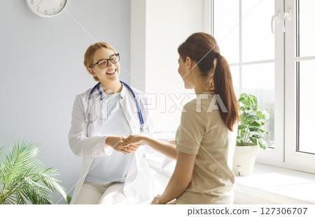 Smiling female family doctor and patient shaking hands in clinic after routine health consultation. Smiling female family doctor and patient shaking hands in clinic after routine health consultation. 127306707