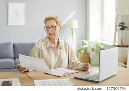 Woman with glasses reviewing documents and writing notes at desk, working from home in bright room. Woman with glasses reviewing documents and writing notes at desk, working from home in bright room. 127306764