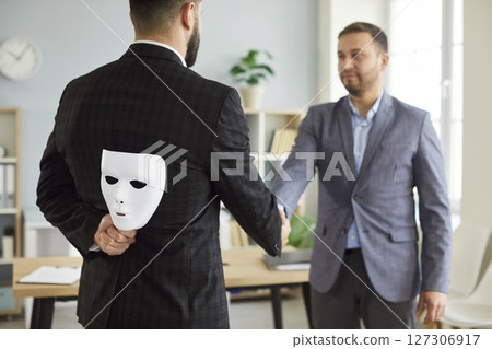 Businessman wearing suit giving dishonest handshake hiding white face mask behind his back. 127306917