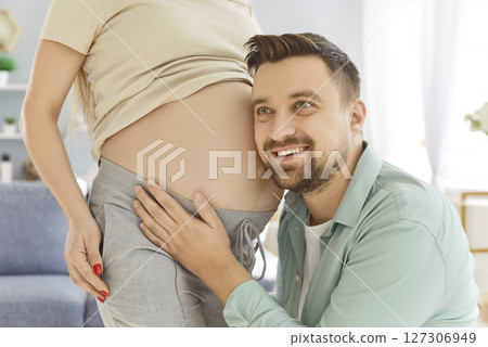 Happy smiling excited expectant father cuddling his wife pregnant belly, listening to baby moving. Happy smiling excited expectant father cuddling his wife pregnant belly, listening to baby moving. 127306949
