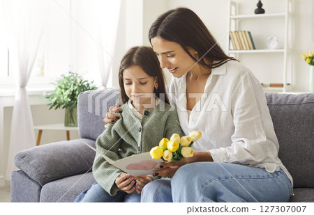 Young mother and daughter sitting on sofa at home and reading a greeting card together. Young mother and daughter sitting on sofa at home and reading a greeting card together. 127307007