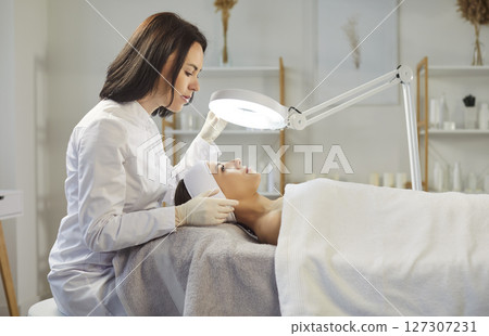 Professional female dermatologist cosmetologist, beautician doing female face exam study under lamp Professional female dermatologist cosmetologist, beautician doing female face exam study under lamp 127307231