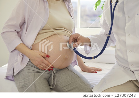 Pregnant woman receiving prenatal checkup in clinic while doctor examining belly with stethoscope. 127307248