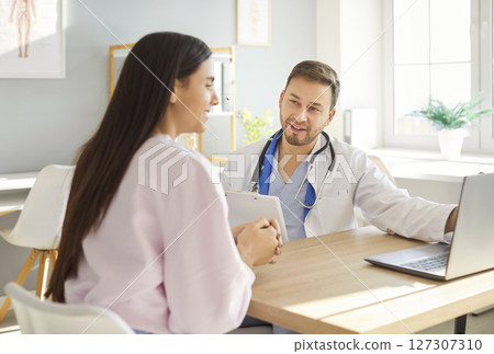 Smiling male doctor in white medical uniform meets to show checkup results on laptop, consult female Smiling male doctor in white medical uniform meets to show checkup results on laptop, consult female 127307310