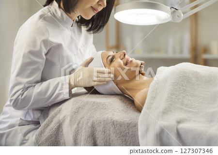 Woman cosmetologist exam young woman skin condition under lamp in beauty spa salon. Woman cosmetologist exam young woman skin condition under lamp in beauty spa salon. 127307346