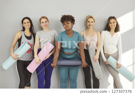 Diverse happy female sporty women group standing in a row in gym holding yoga mat 127307376