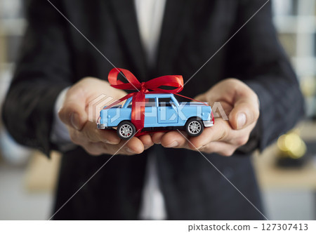 Salesman in suit holding blue toy car with red bow symbolizing buying vehicle or auto insurance. 127307413